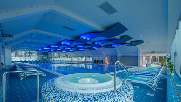 An indoor swimming pool with a central hot tub, blue tiled floor, and a ceiling with blue lighting. - Hotel Cerkno (Cerkno)