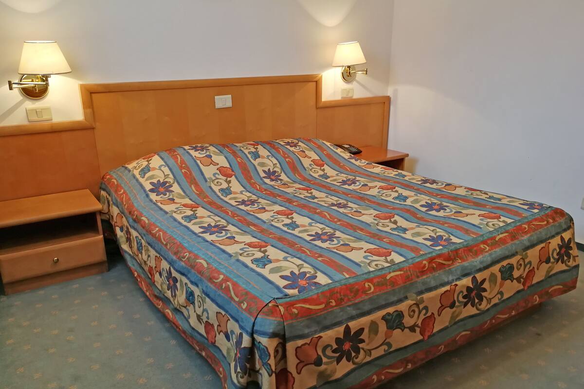 A bed with a floral-patterned bedspread, two bedside lamps, a wooden headboard, and a small wooden nightstand with a drawer.