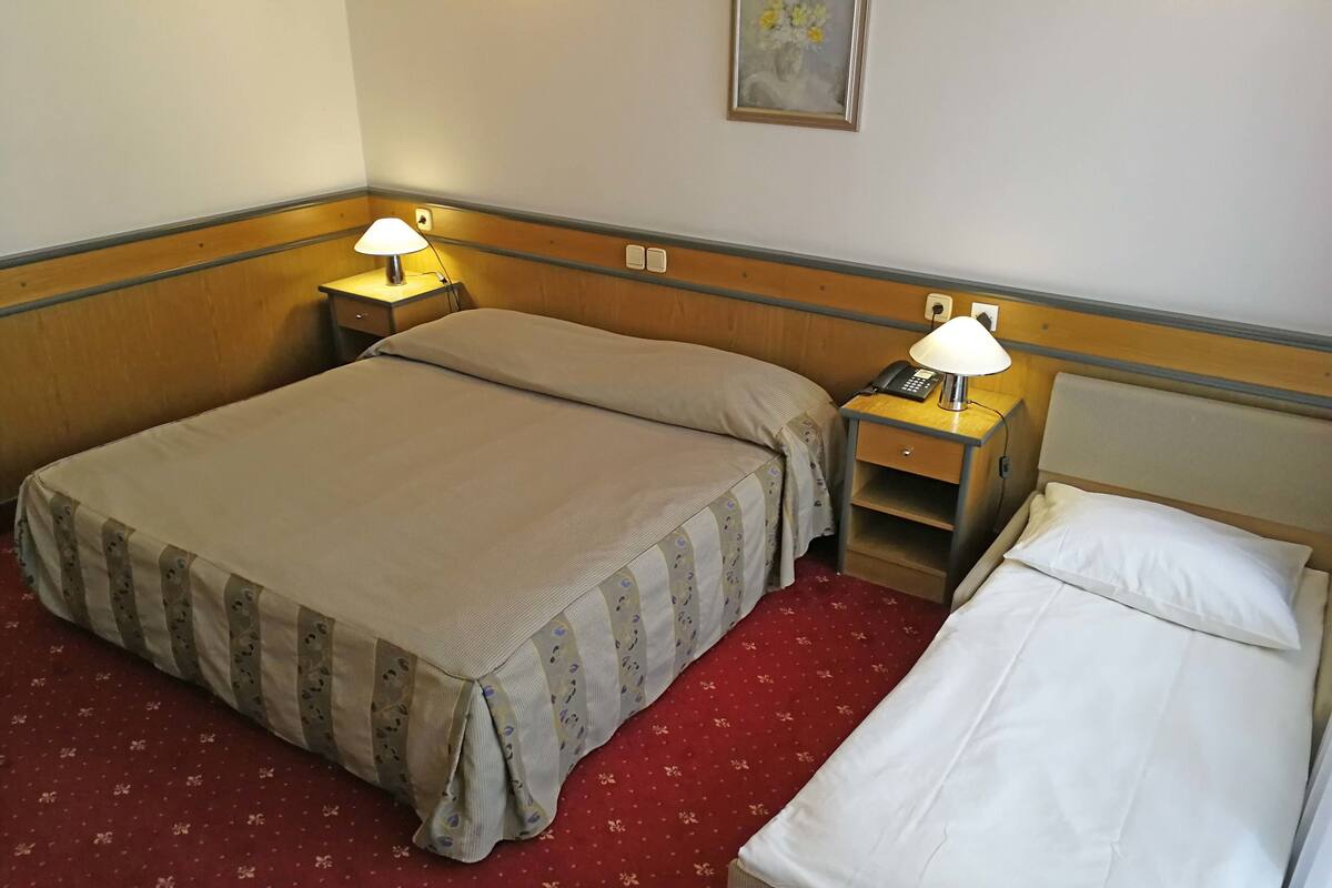 A hotel room with a double bed, a single bed, a nightstand with a lamp, and a telephone.