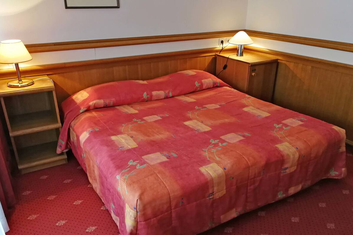 A neatly made bed with a red and orange patterned bedspread, two bedside lamps, and a wooden nightstand with a shelf.