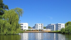 Ein modernes Gebäude mit flachem Dach und weißen Außenwänden, neben einem ruhigen Gewässer, im Vordergrund ein Weeping Willow Baum.