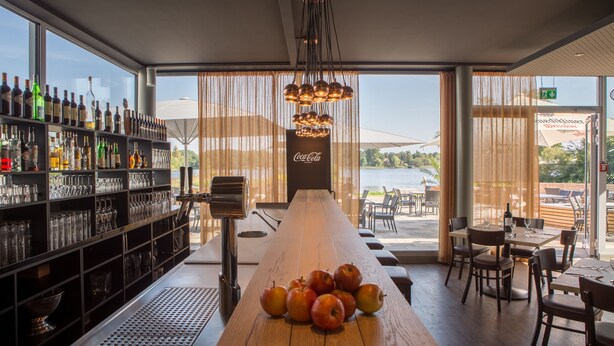 Eine Bar mit einem langen Holztisch, Regalen voller Flaschen und Gläsern sowie Blick ins Freie.