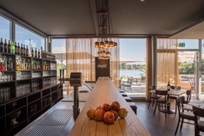 A bar area with a long wooden table, shelves with bottles and glasses, and a view of the outdoors.