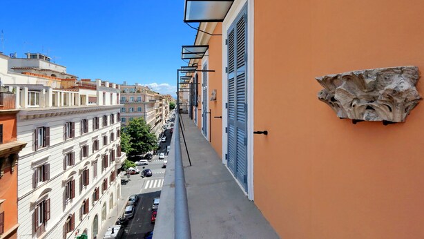 A narrow balcony with a view of a street lined with buildings, a stone wall feature, and a clear blue sky. - Luxurious three-room apartment with balcony, at the corner of Via Veneto (Roma)