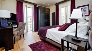 A modern hotel room with a large bed, a desk, and a chair. There is a window with curtains, a radiator, and a lamp on a bedside table. - Luxurious three-room apartment with balcony, at the corner of Via Veneto (Roma)