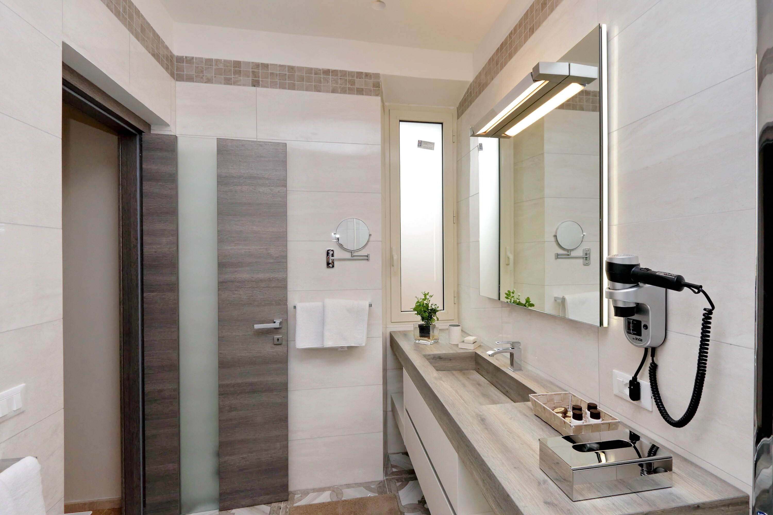 A modern bathroom with a glass shower enclosure, a large mirror, a hairdryer, and a vanity with a sink and storage.