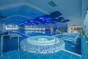 Ein Hallenbad mit einer zentralen Whirlwanne, blau gefliestem Boden und einer Decke mit blauer Beleuchtung.