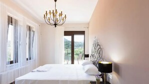 A bedroom with a bed, a bedside table with a lamp, a window with curtains, and a view of the outdoors. - Eudora Luxury Stone Villa (Apokoronas)