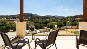 A balcony with a glass railing, two chairs, a small table with a cup and a plant, and a view of a hilly landscape. - Eudora Luxury Stone Villa (Apokoronas)