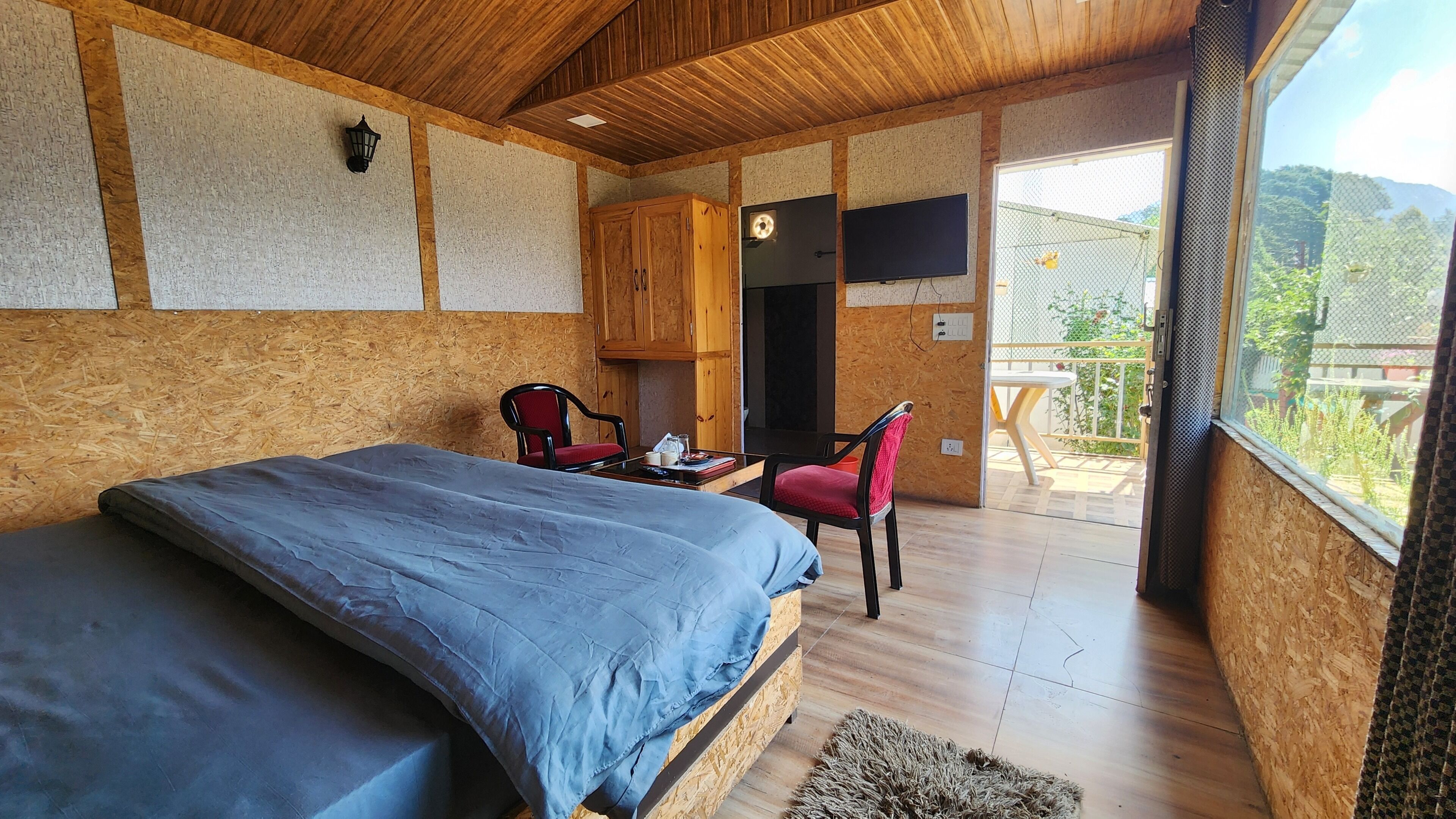Ein Schlafzimmer mit einem Bett, einem kleinen Tisch und einem Stuhl. Eine Holzbalkendecke und ein Blick ins Freie durch ein Fenster.
