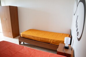 Uma cama de solteiro com estrutura de madeira e colchão, um guarda-roupa de madeira, uma mesa de cabeceira com abajur e um tapete vermelho.