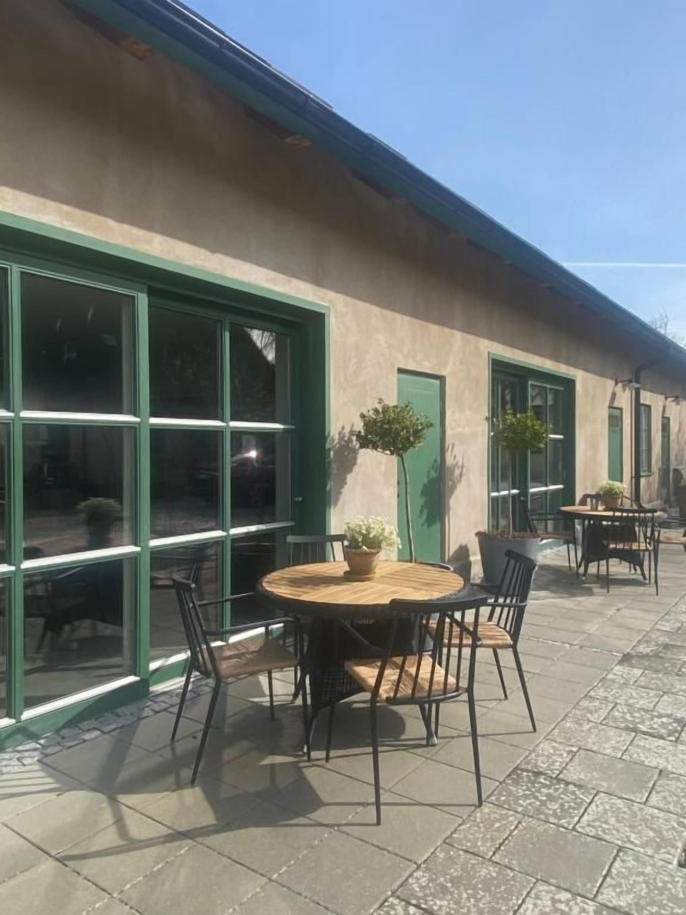 Een terras met een ronde tafel en stoelen, een plant in een pot, en een gebouw met grote ramen en een groene deur.