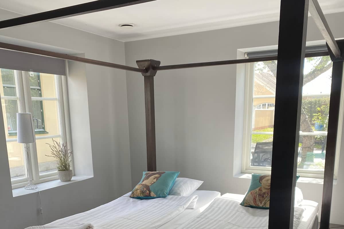 Uma cama de dossel com lençóis brancos e duas almofadas, uma planta em vaso e uma janela com vista para o exterior.