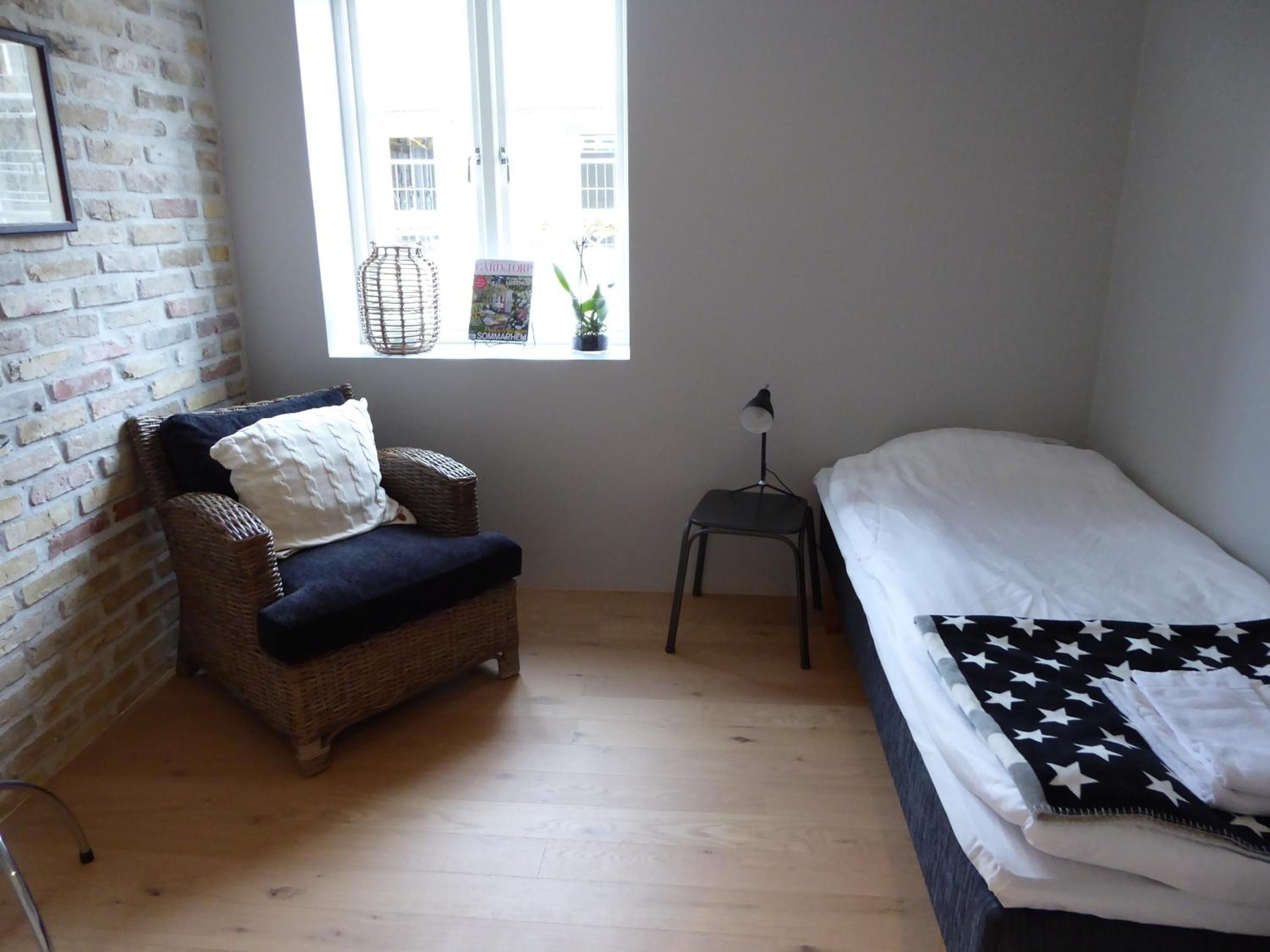 A room with a brick wall, a wicker chair, a small table with a lamp, a bed with a star-patterned blanket, and a window with a view of buildings.