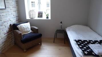 A room with a brick wall, a wicker chair, a small table with a lamp, a bed with a star-patterned blanket, and a window with a view of buildings.