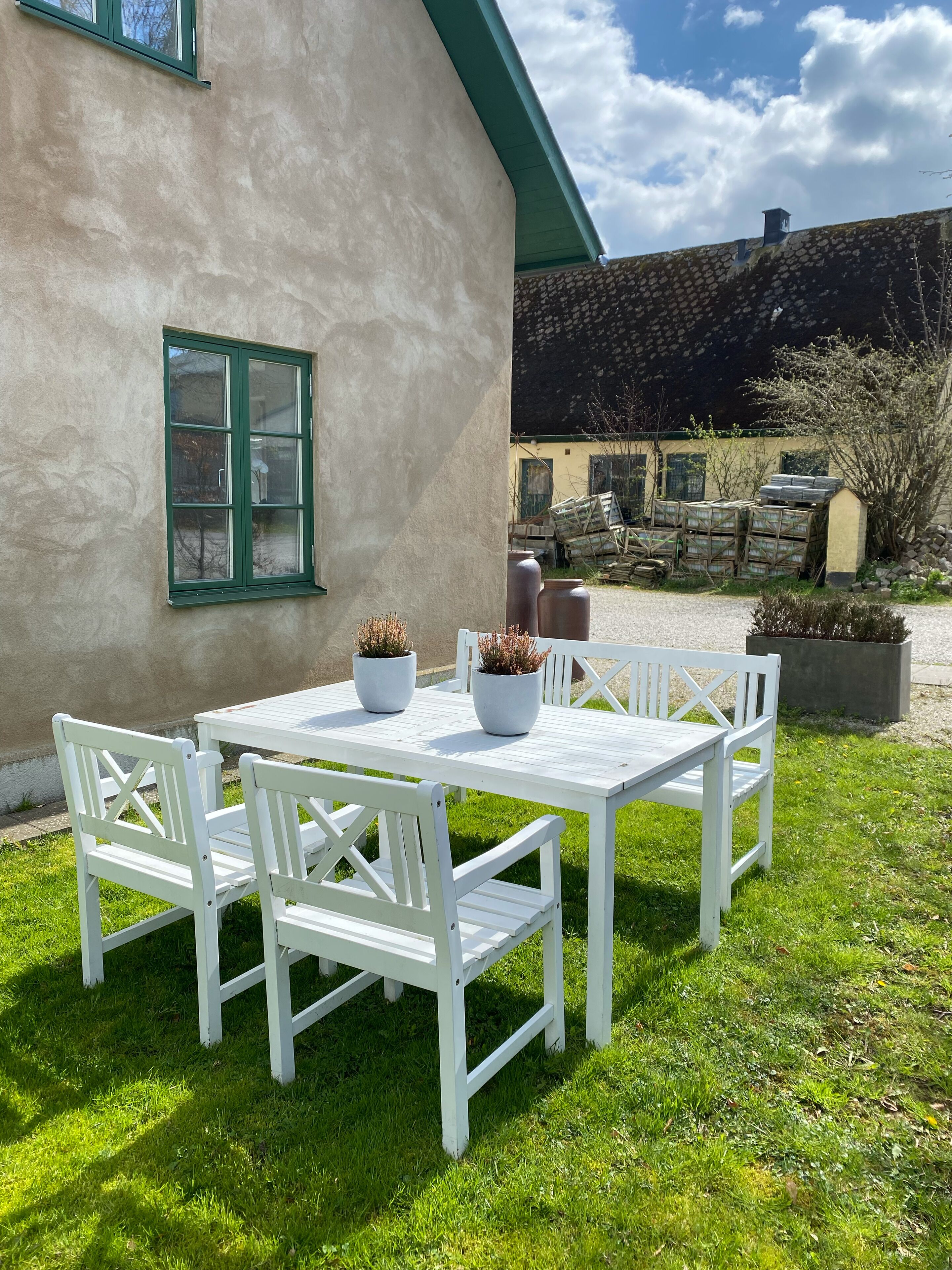 A white outdoor table with chairs, potted plants, and a window with green shutters.