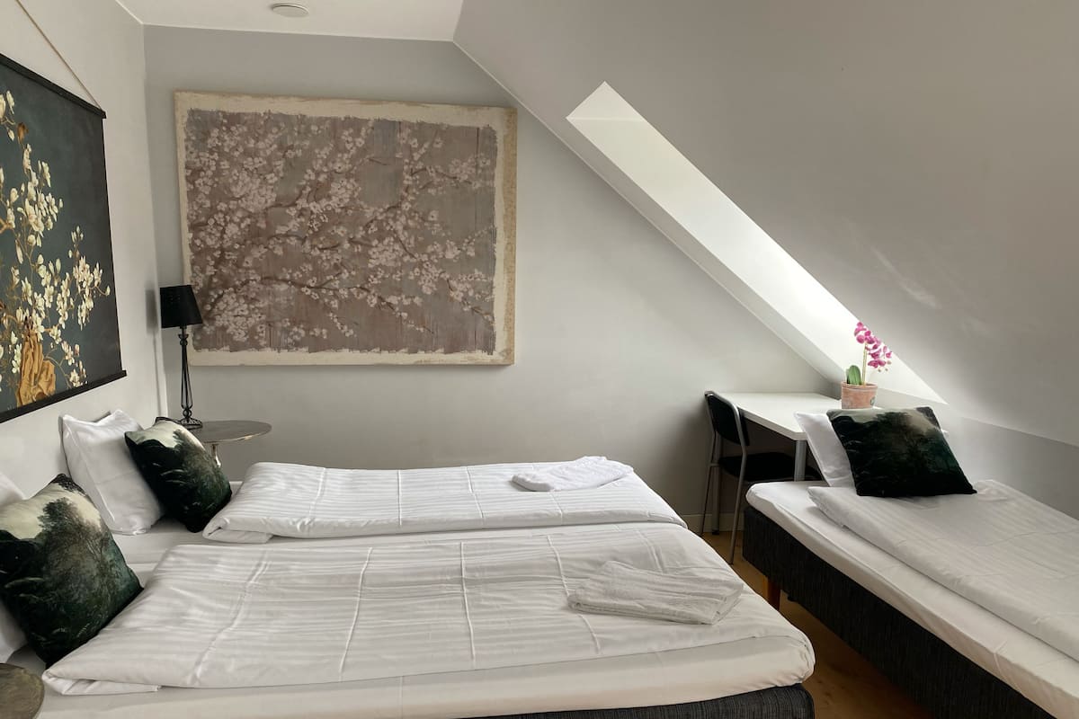 Quarto moderno com duas camas, uma grande obra de arte floral, uma mesa pequena e um vaso com planta.