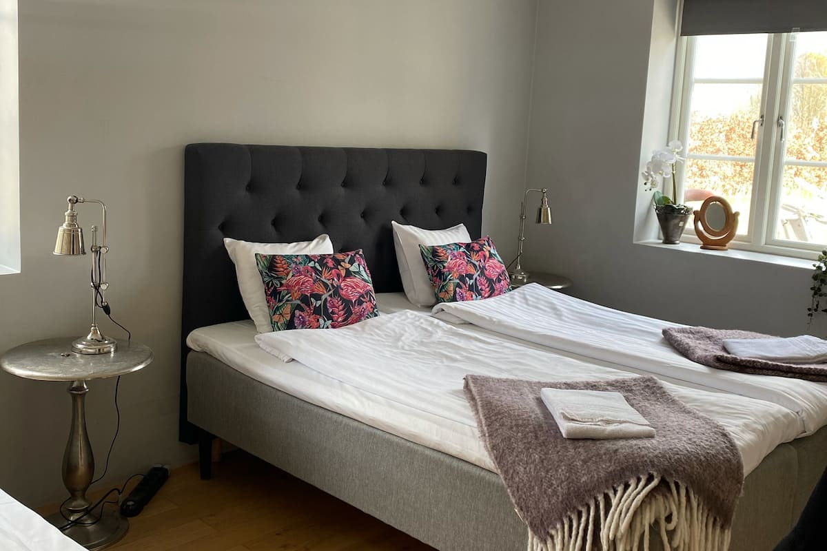 Quarto com uma cama grande, uma mesa de cabeceira, uma luminária e uma janela com vista para árvores.