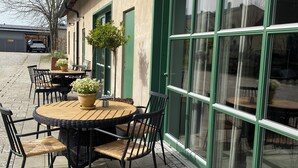 Un patio al aire libre con una mesa redonda y sillas, una planta en maceta y un edificio con ventanas amplias y contraventanas verdes.