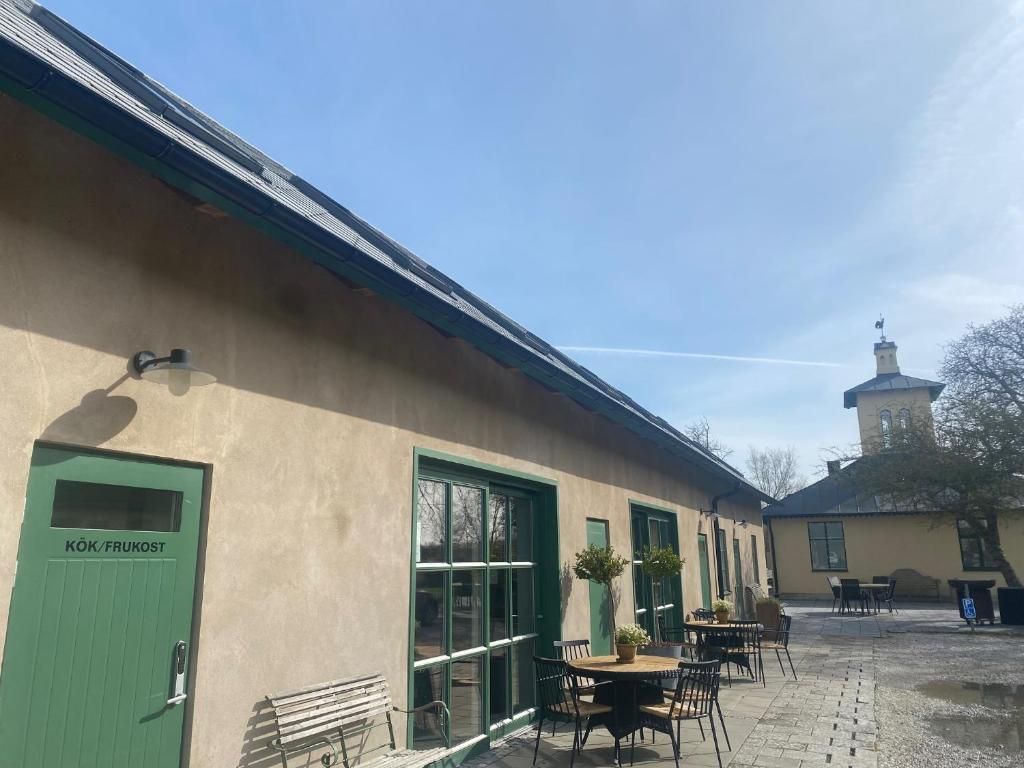 A building with a green door labeled 'KÖK/FRUKOST,' outdoor seating with tables and chairs, and a clear sky.