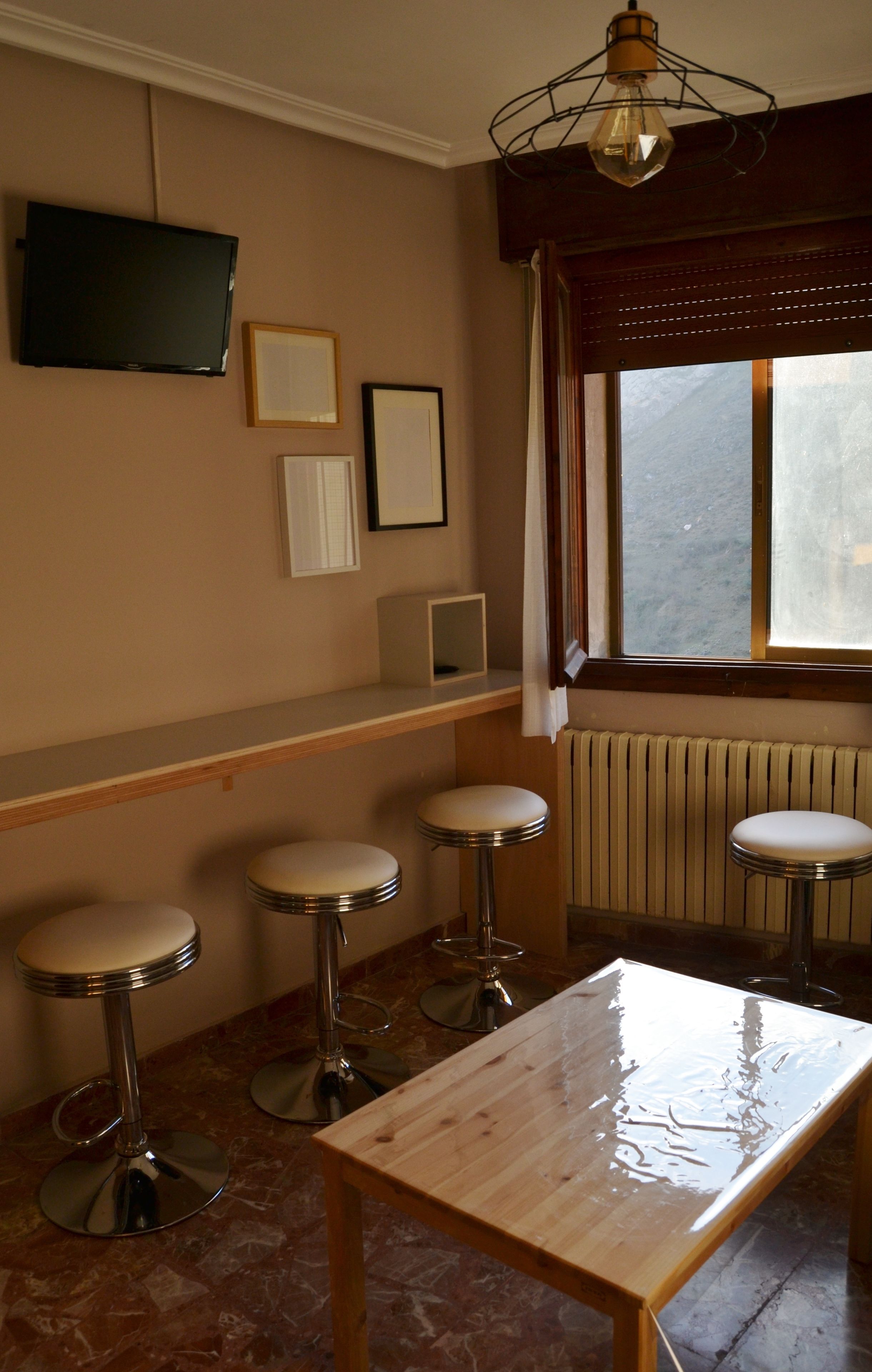 Ein Zimmer mit einem Holztisch, vier Barhocker, einem an der Wand montierten Fernseher und einem Fenster mit Vorhängen.