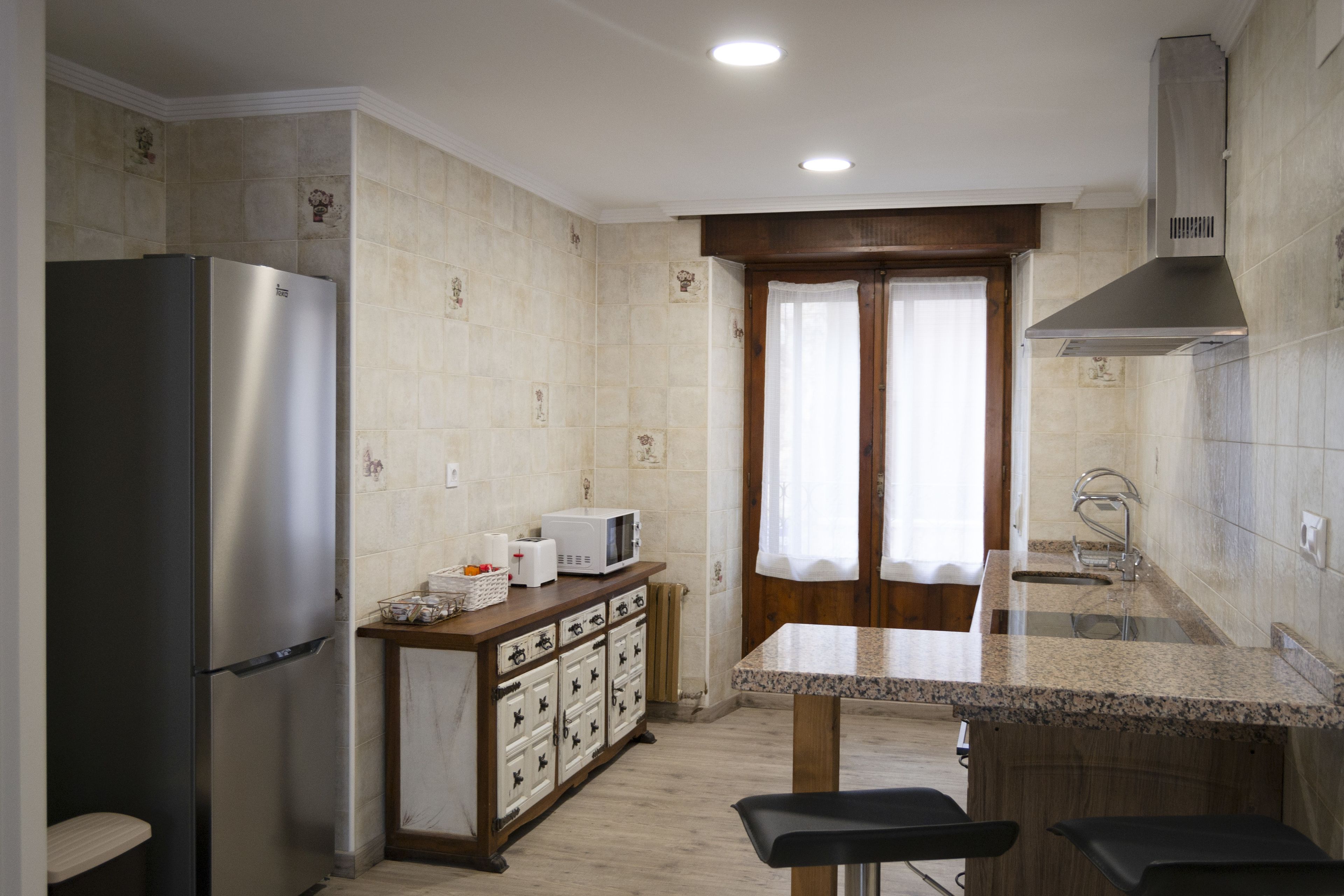 Una cocina con refrigerador, microondas, gabinetes de madera, un lavamanos y una ventana con cortinas.