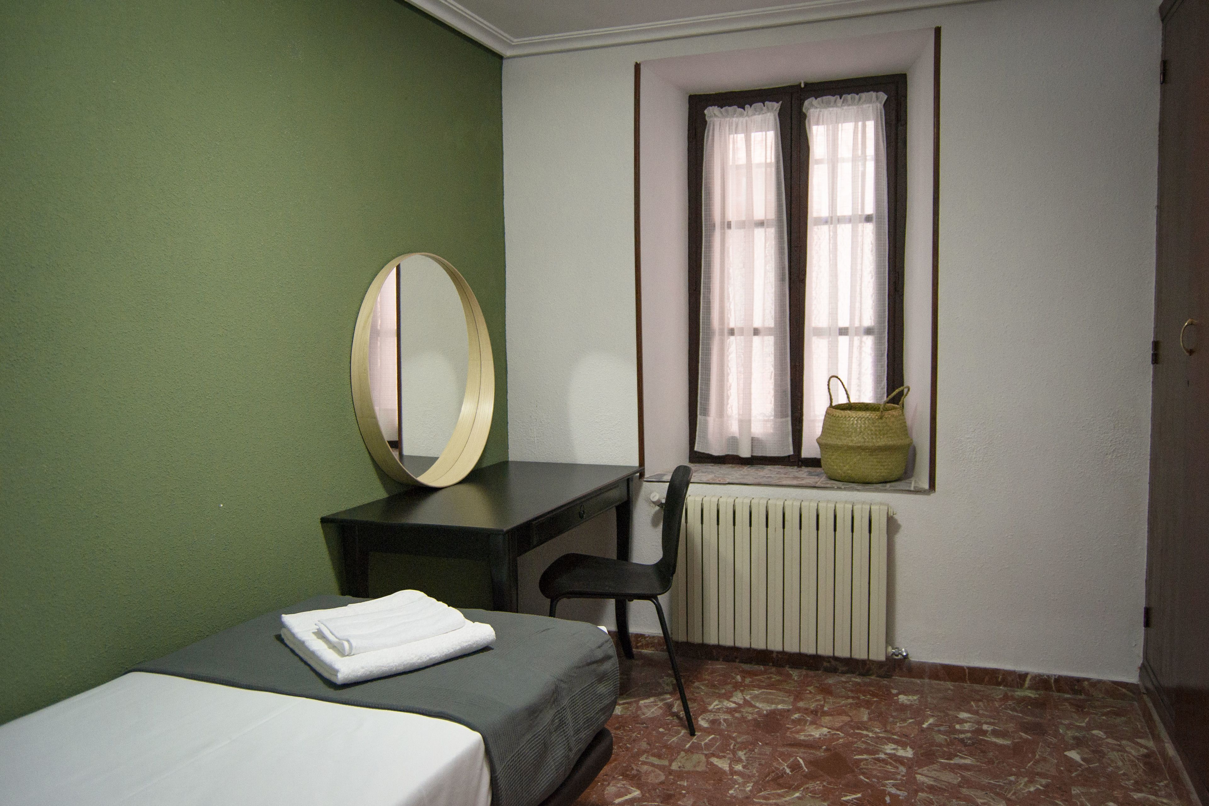 A small, simple room with a bed, a desk, a chair, a large mirror, and a window with a curtain.
