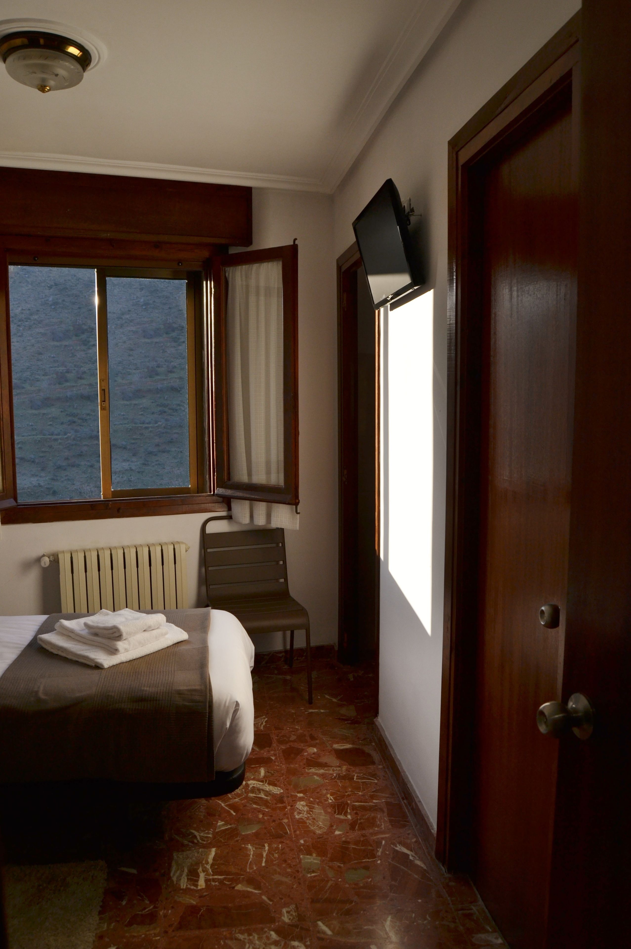 A hotel room with a bed, a chair, a TV mounted on the wall, and a window with curtains.