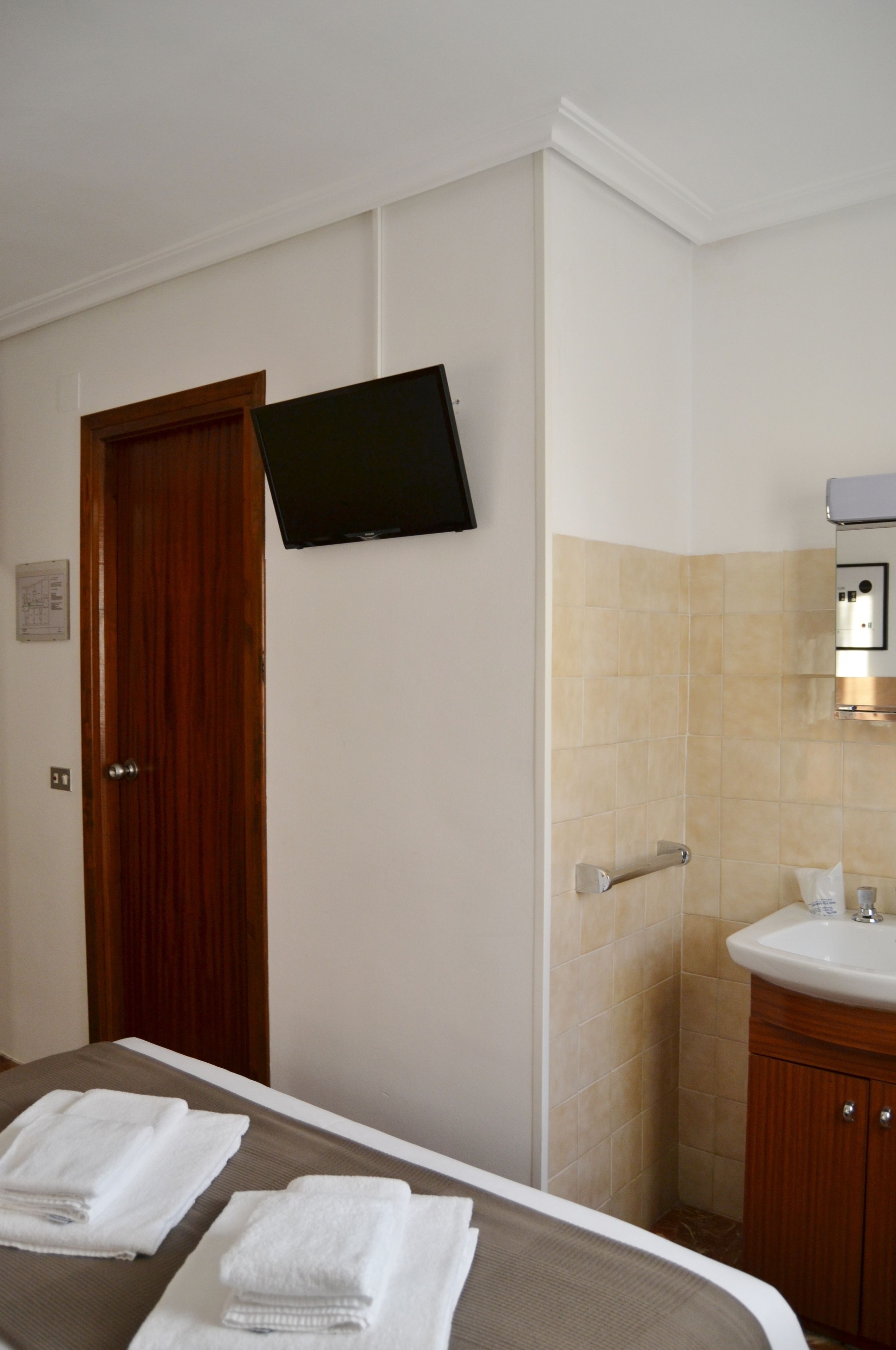 Een hotelkamer met een bed, een aan de muur bevestigde televisie, een badkamer met een wastafel en spiegel, en een deur.