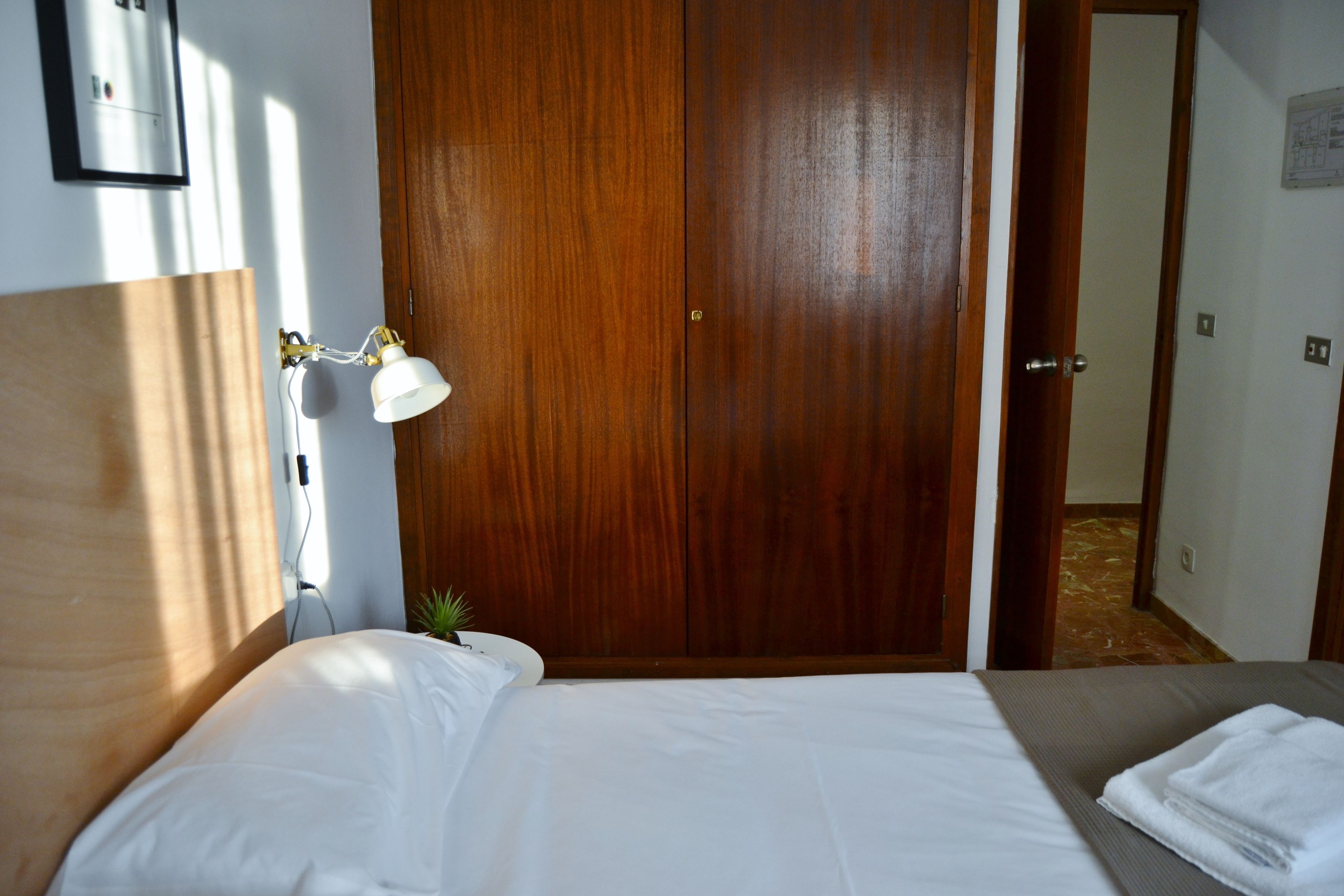 Une chambre d’hôtel avec une armoire en bois, une lampe de chevet, un lit avec du linge de lit blanc et une petite plante en pot.