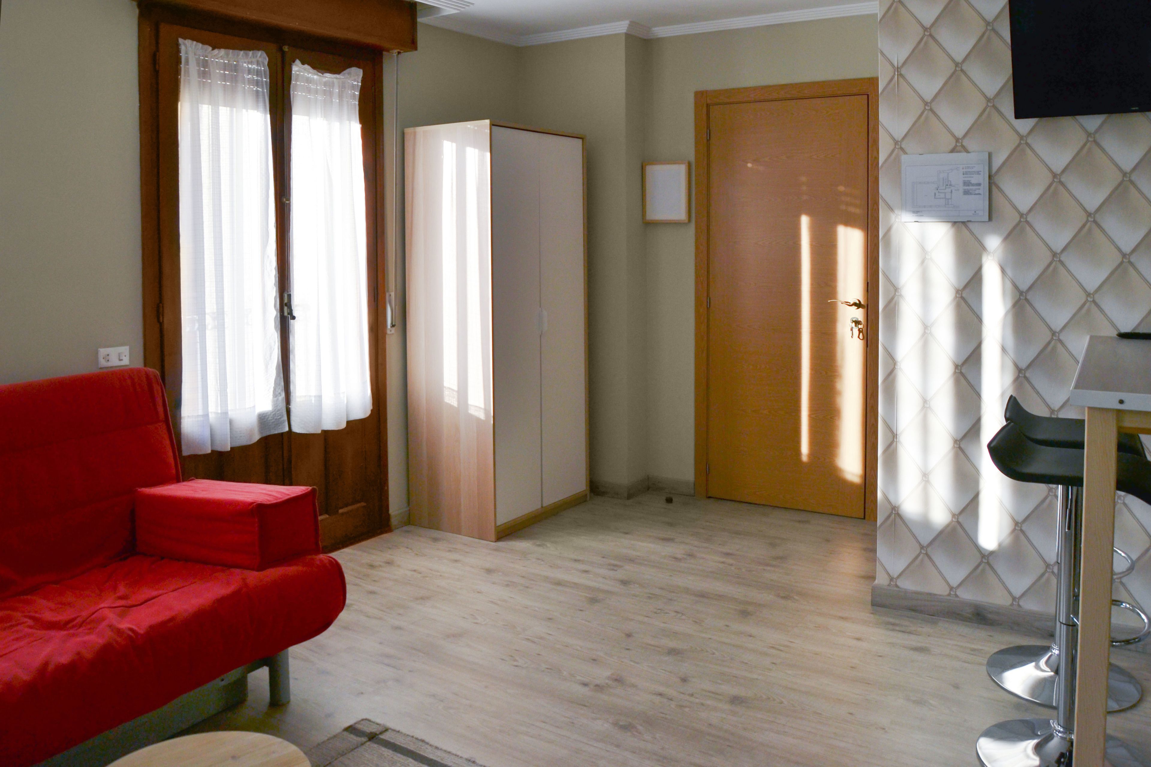 Una habitación con un sofá rojo, un armario, una puerta y una pequeña mesa con una silla.