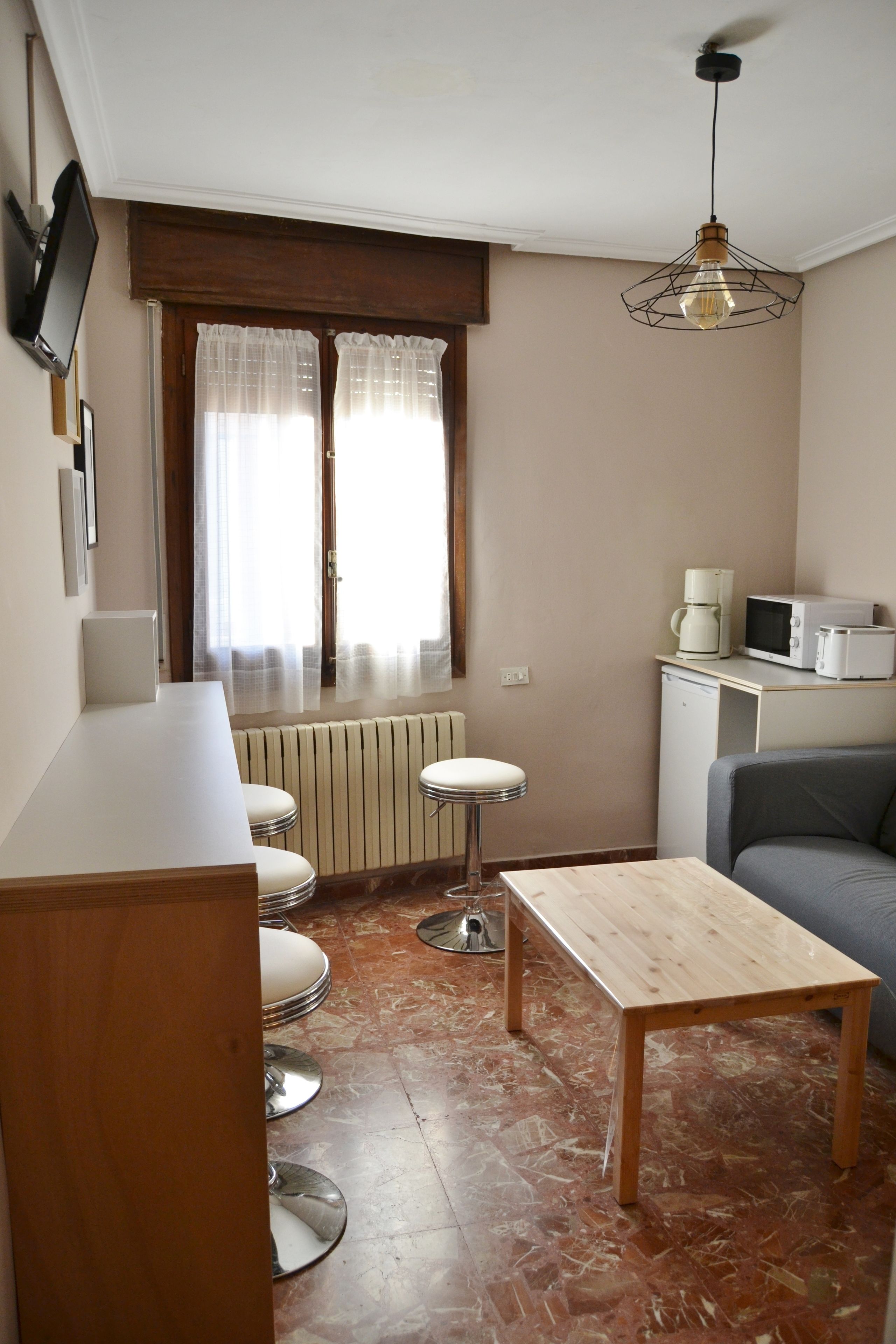 Un espace de vie compact comprenant une table à manger, deux tabourets, un canapé, une petite kitchenette et une fenêtre avec des rideaux.