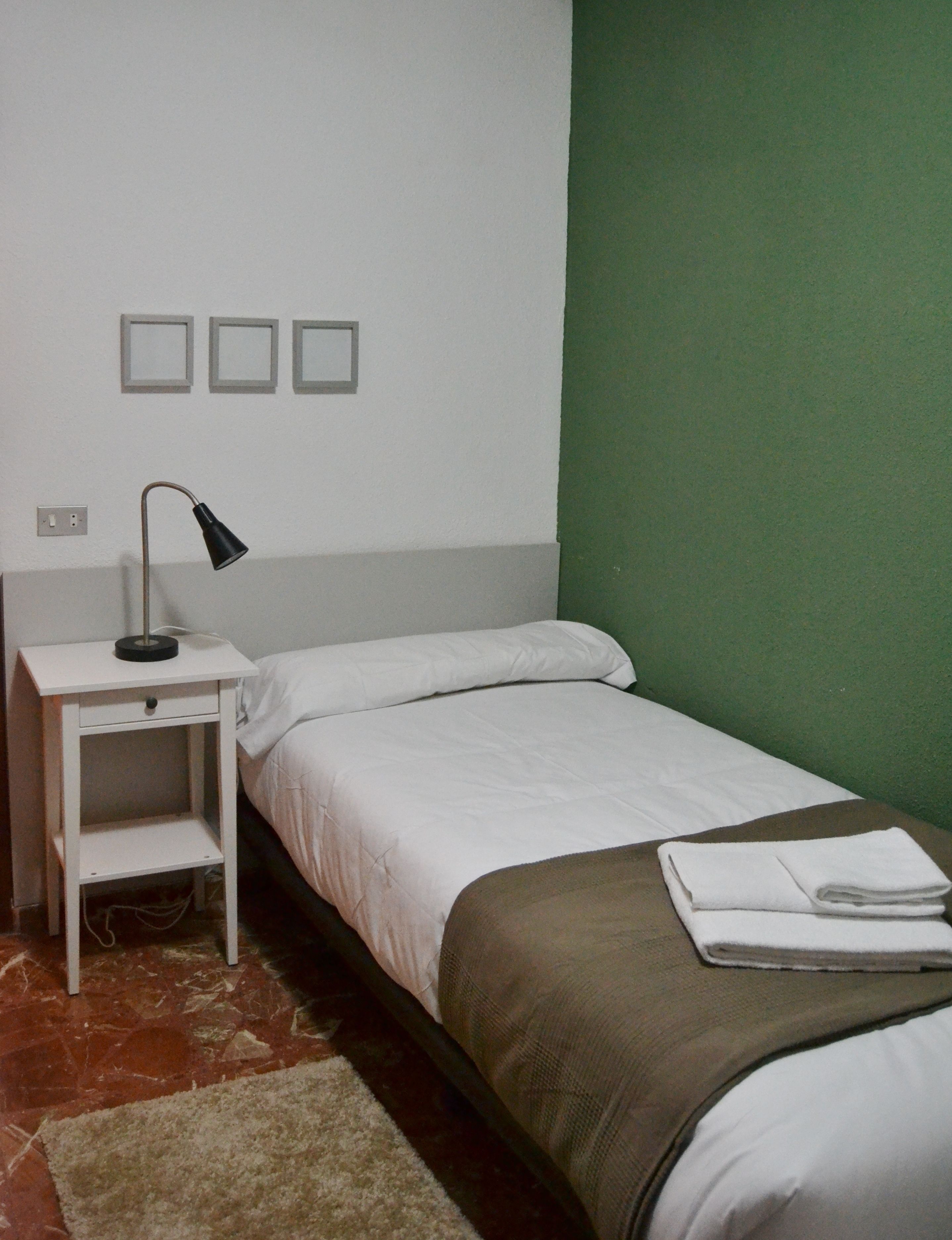 Una cama individual con ropa de cama blanca, una mesita de noche con lámpara y una pared verde.