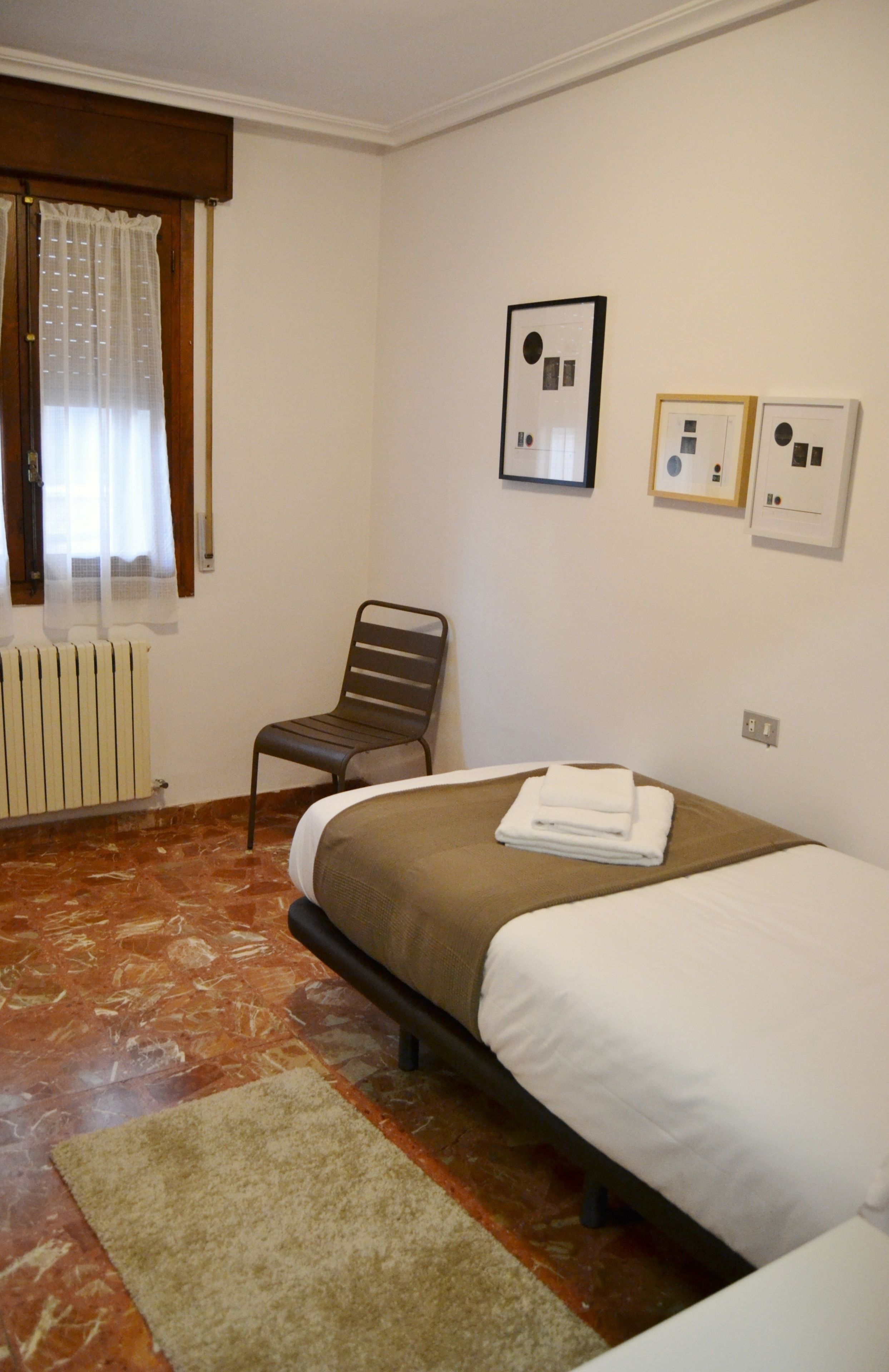 A hotel room with a bed, a chair, a radiator, and a window with curtains.