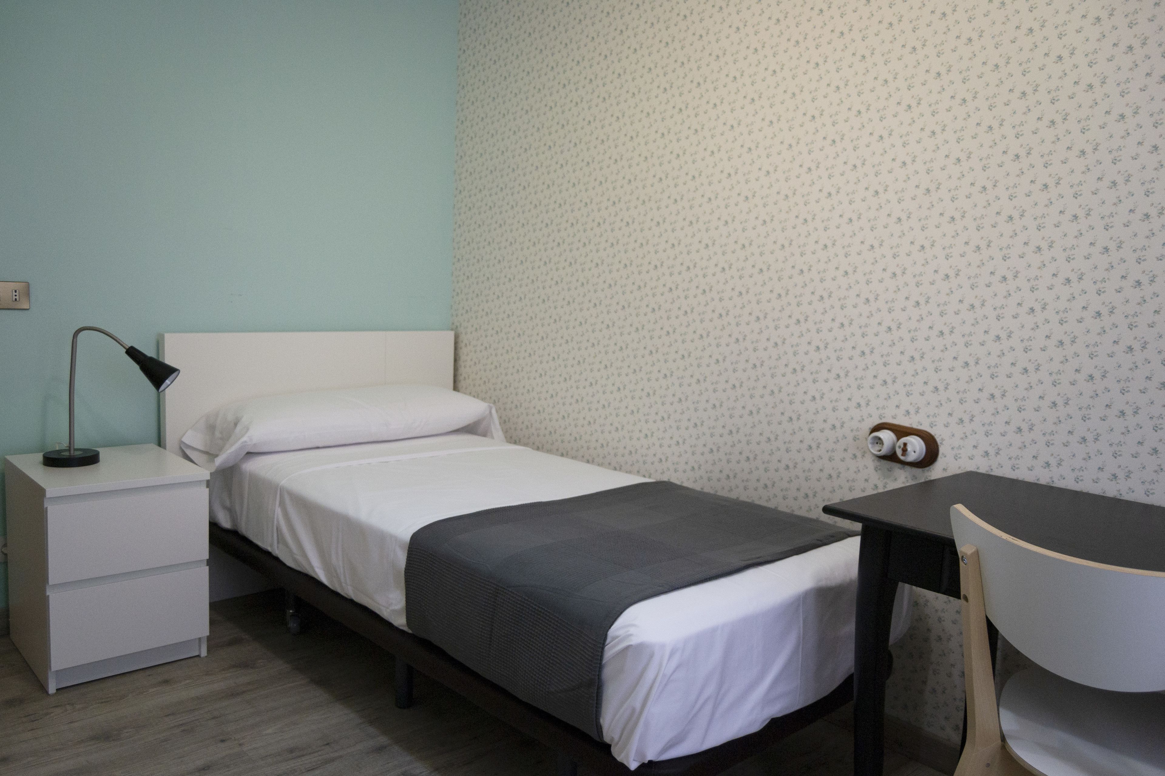 Een kleine, verzorgde slaapkamer met een enkel bed, een nachtkastje met lamp, een stoel en een bureau.