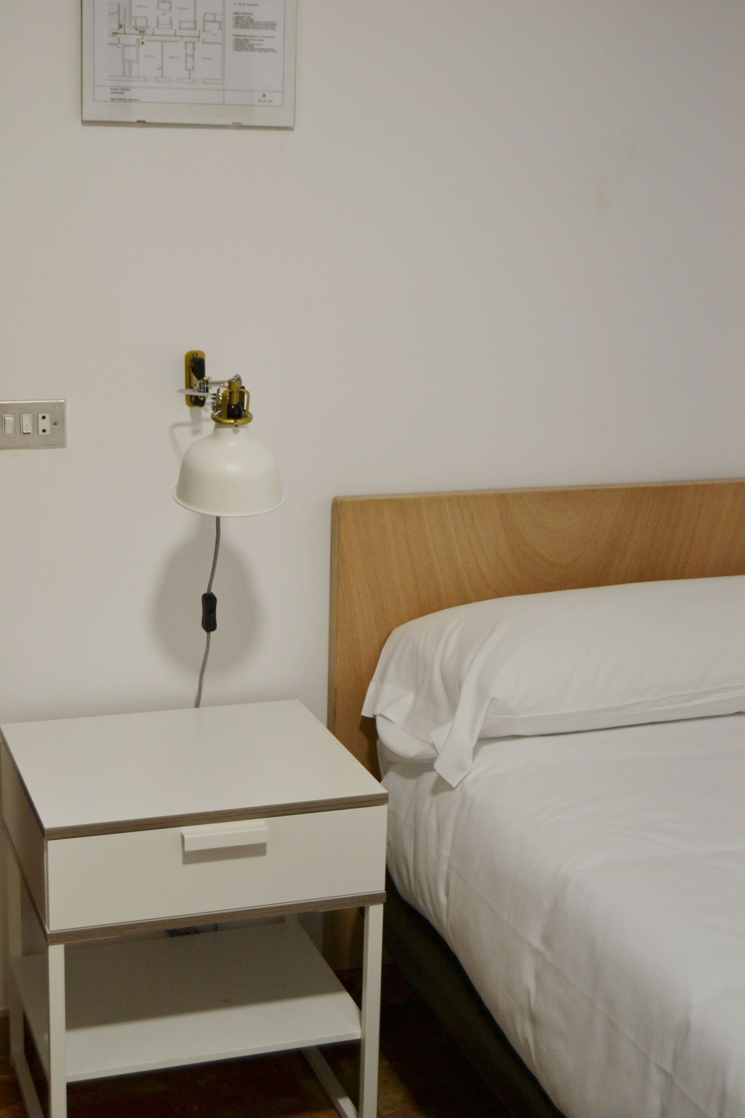 Quarto simples com cama, mesa de cabeceira com abajur e uma tomada na parede.