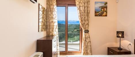A bedroom with a bed, a dresser, a nightstand, a lamp, a painting, and a view of mountains through a sliding door.