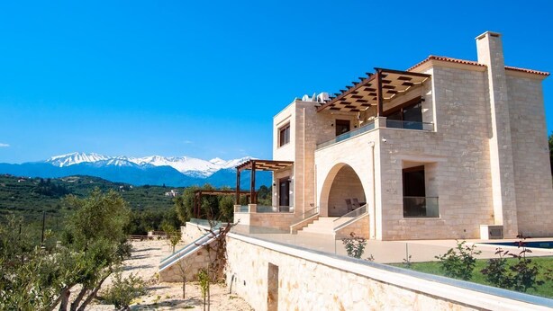 A modern two-story house with a stone facade, a covered patio, and a view of snow-capped mountains.