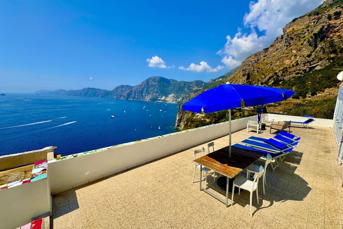 Une terrasse avec une table et des chaises, un parasol bleu, et une vue sur la mer et les montagnes.