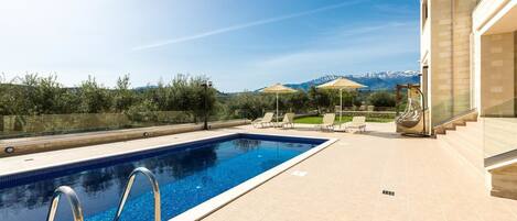 A swimming pool with clear blue water, surrounded by beige tiles. There are lounge chairs and umbrellas on the patio area.