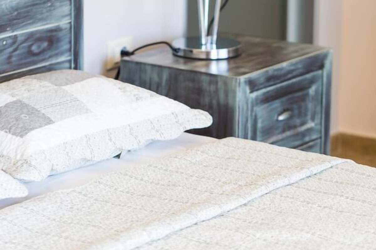 Cama bem arrumada com colcha branca texturizada e um travesseiro com padrão geométrico. Uma luminária de chão moderna, com base metálica e cúpula cônica, está posicionada à direita da cama.