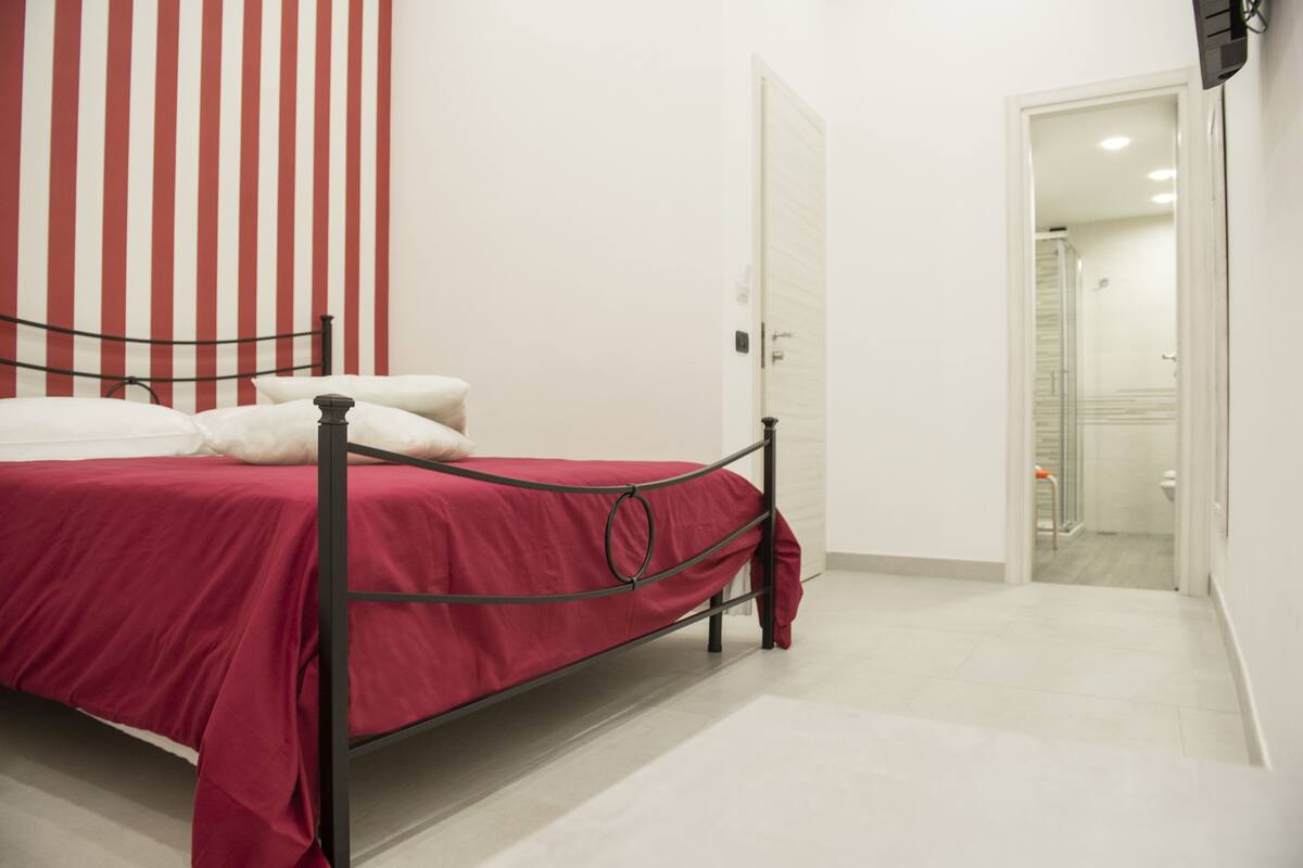 A bedroom with a metal bed frame, a red bedspread, and white pillows. There is a white door and a shower visible in the background.