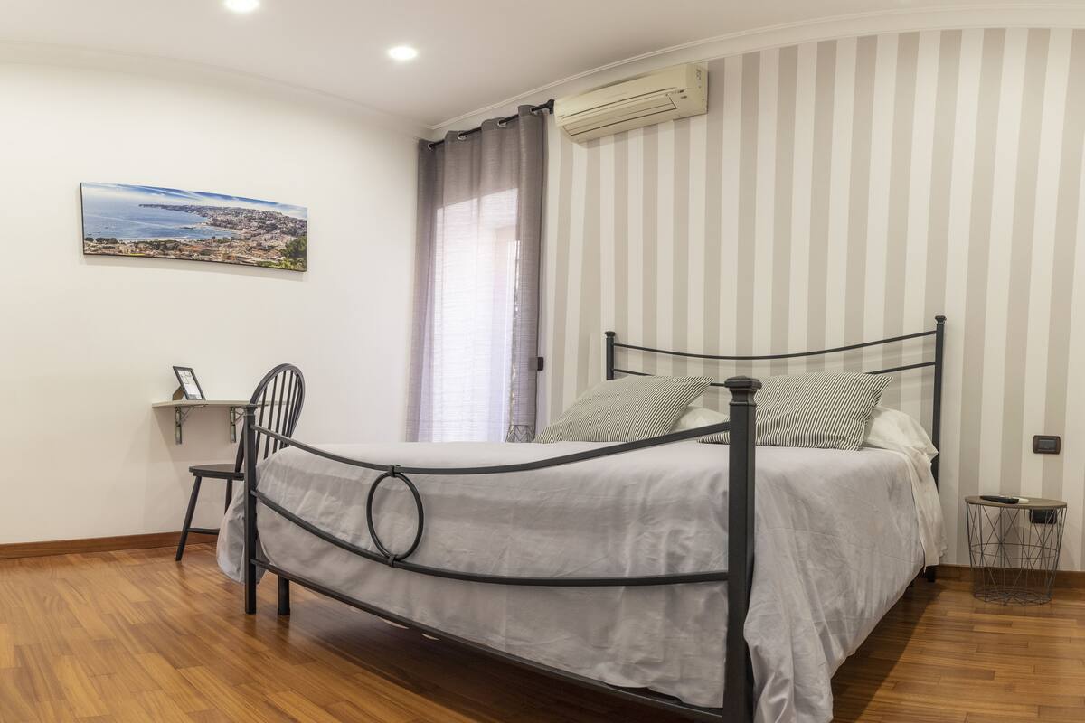 A bedroom with a bed, a chair, a small table, a picture on the wall, and a window with curtains.