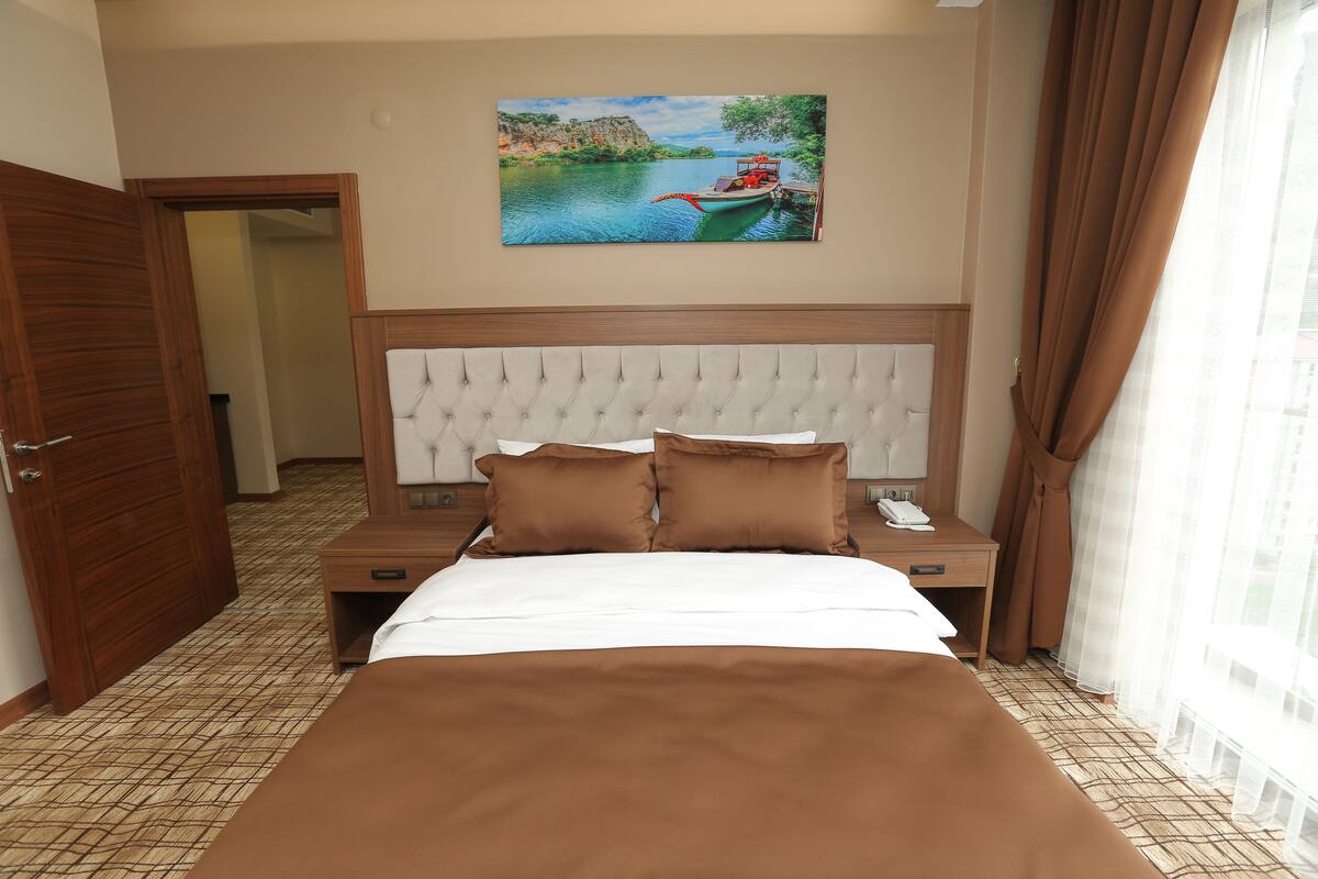 A hotel room with a large bed, a wooden headboard, a painting of a lake scene, and a view of a cityscape through sheer curtains.