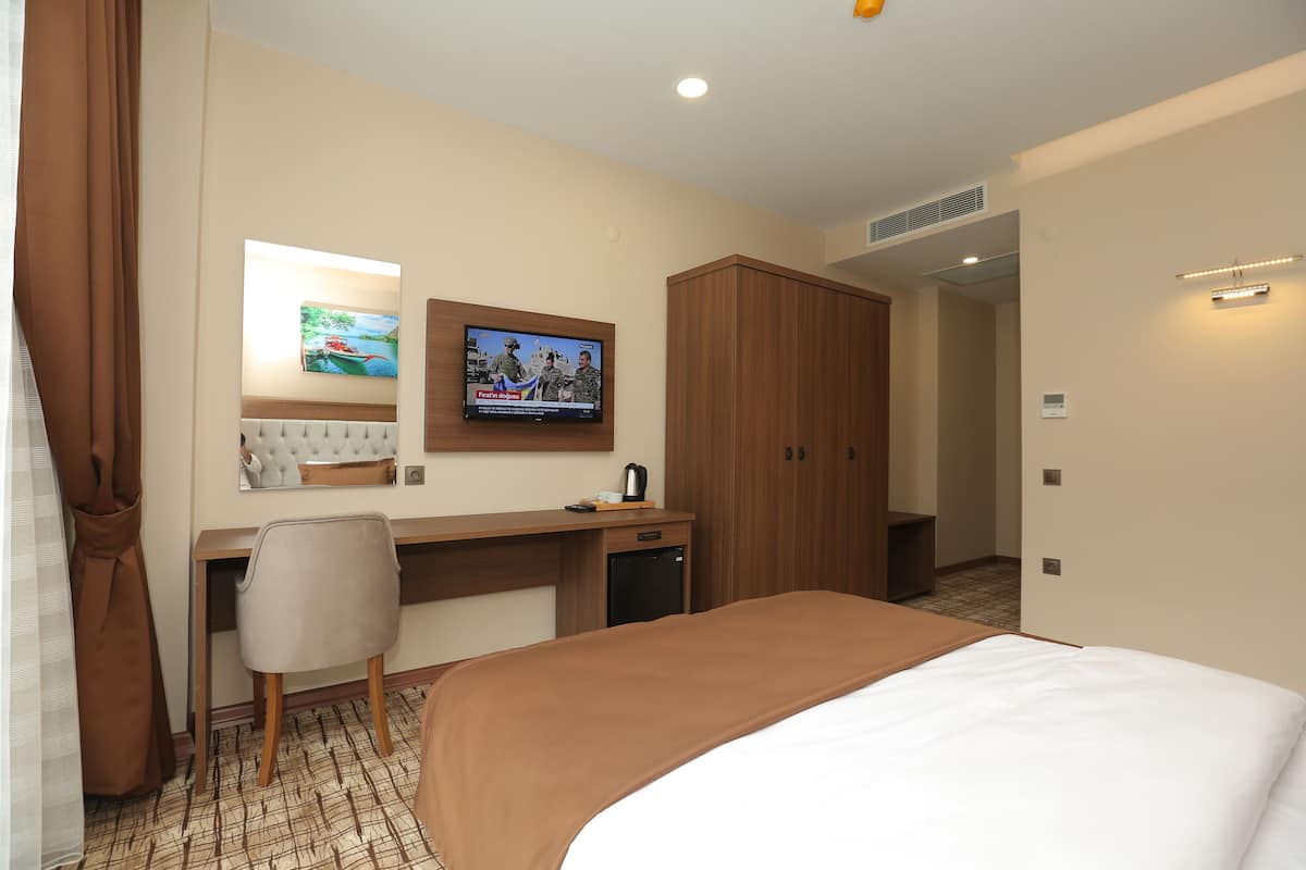A hotel room with a bed, a desk with a chair, a television mounted on the wall, and a wardrobe.