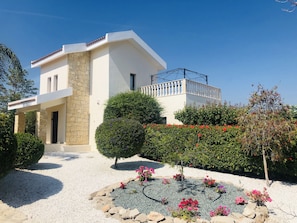 A modern two-story house with a stone facade, a balcony, and a well-maintained garden with various plants and flowers. - A Relaxing, Perfectly located 4 bed Villa in Coral Bay (Peyia)