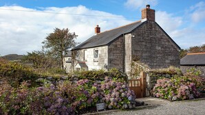 A stone building with a sloped roof and a chimney, surrounded by a gravel driveway and flowering plants. - 3 bedroom accommodation in Nanpean, near St Austell (Nanpean, near St Austell)