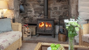 A cozy room with a stone fireplace, wicker furniture, and a table with apples and grapes. - 3 bedroom accommodation in Nanpean, near St Austell (Nanpean, near St Austell)