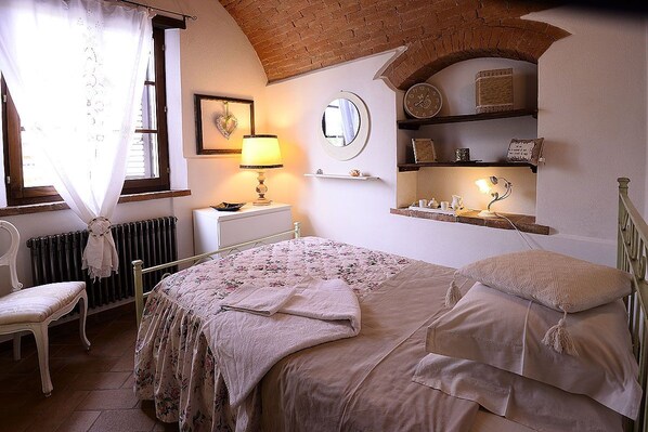 A bedroom with a bed, a chair, a radiator, a shelf with decorative items, and a window with curtains. - old barn on Via Francigena (Siena)