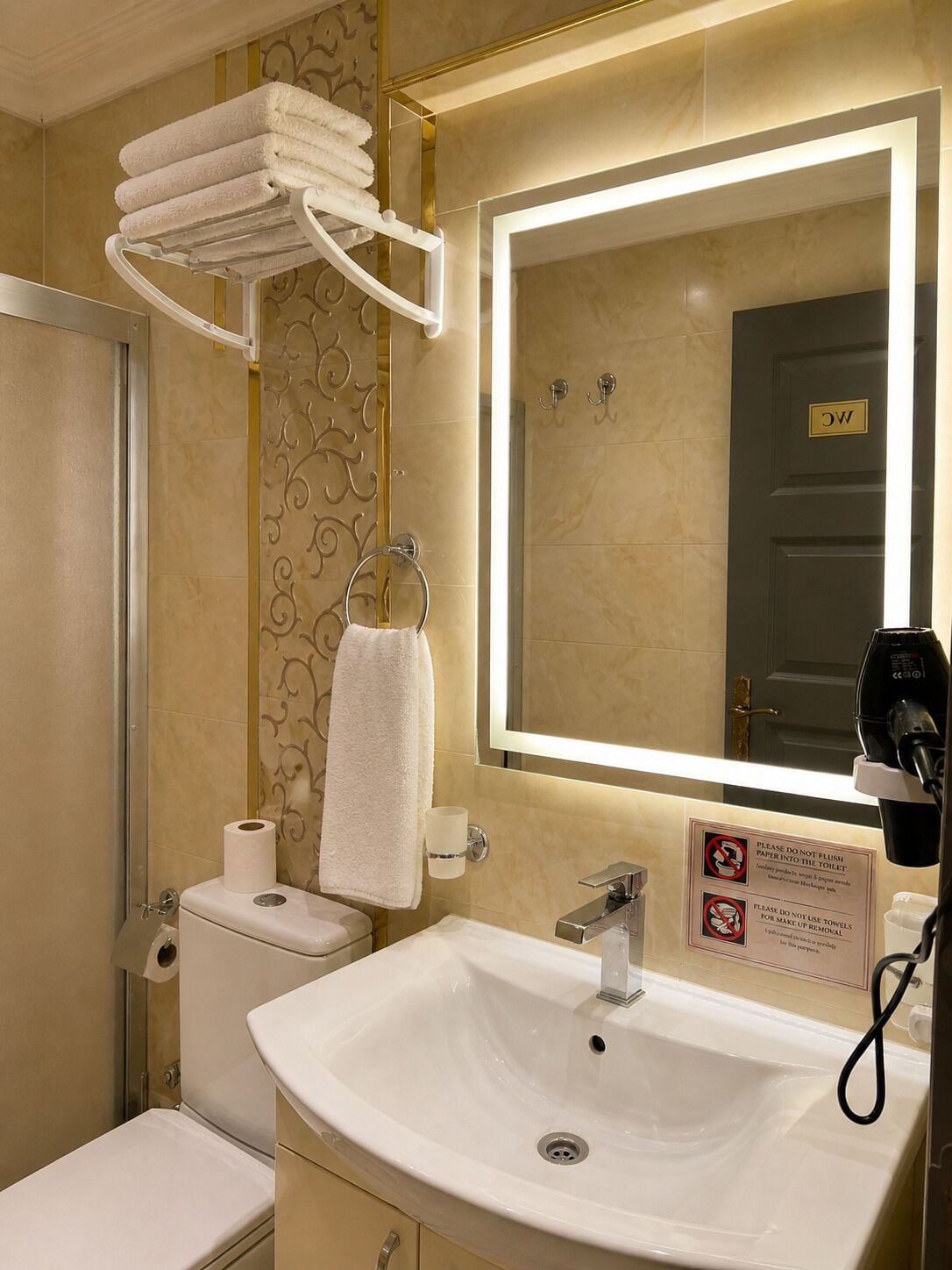 Un baño de hotel con lavamanos, inodoro, toallero y espejo con luz.